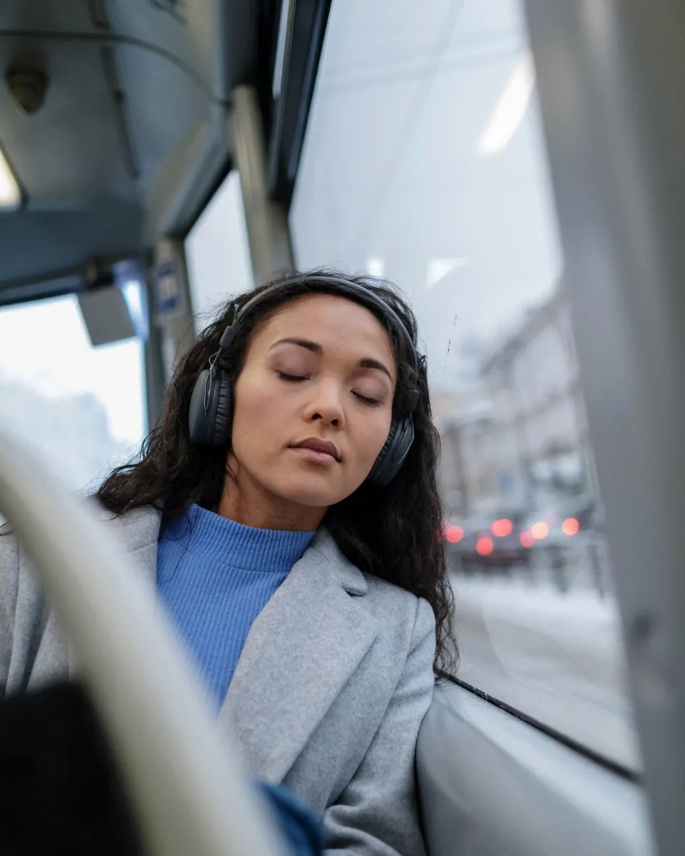 A woman with headphones on resting against a bus window, eyes closed