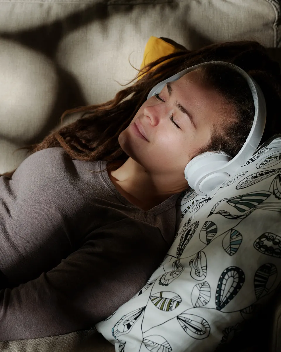 A woman lying on a couch in dappled evening light with headphones on, eyes closed