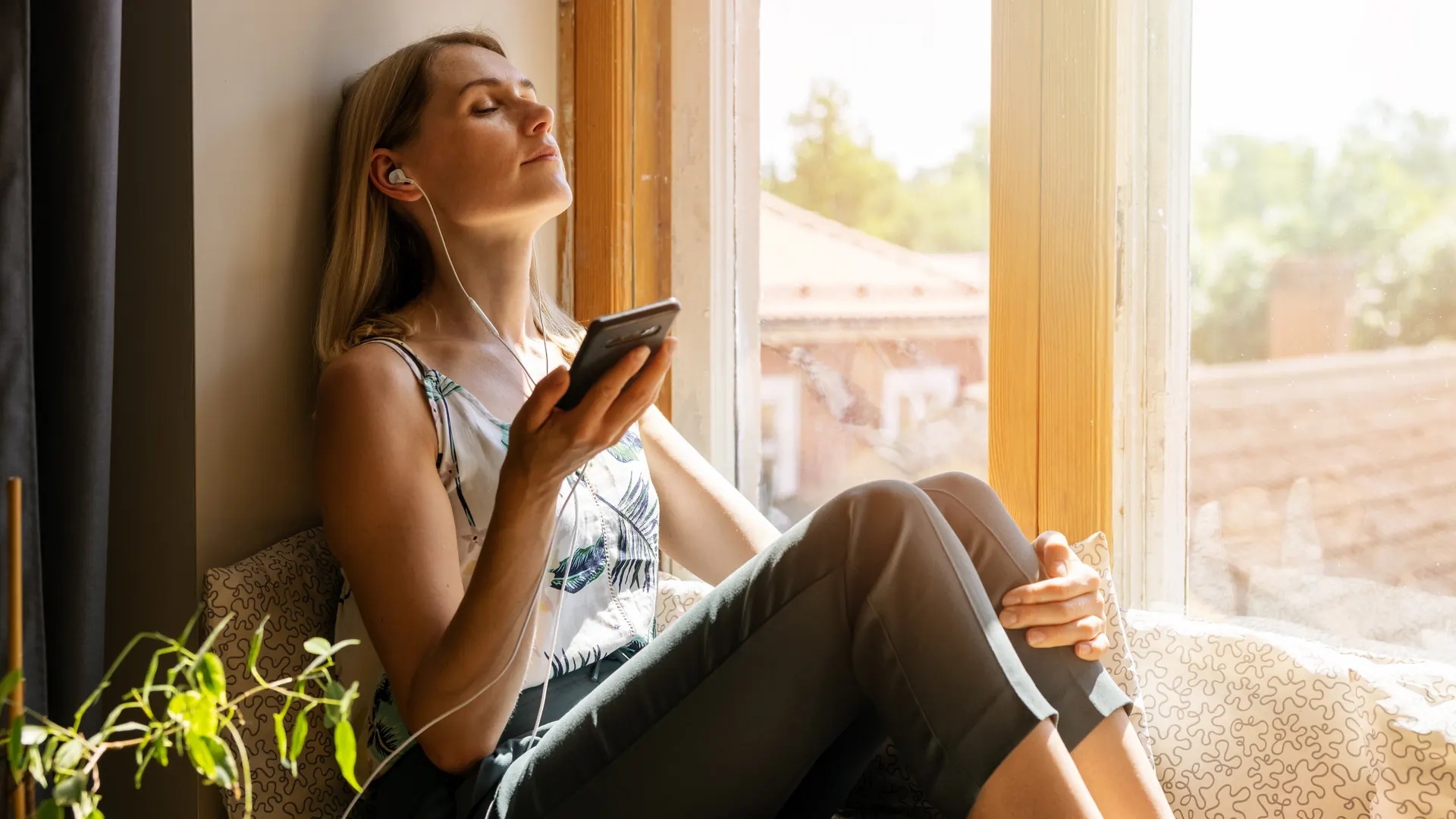 A woman sitting on a sunny windowsill in the morning with earphones in, eyes closed
